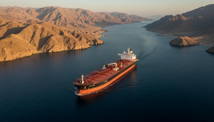 Огромный нефтяной танкер плывет по узкому Ормузскому проливу на фоне скалистых, засушливых берегов.