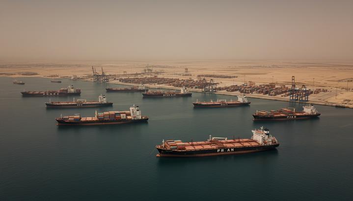 Крупные торговые суда и танкеры стоят на якоре в море на фоне иранского портового города и засушливого побережья.