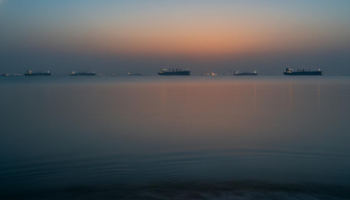 Морская гладь Ормузского пролива с силуэтами грузовых судов на горизонте в вечерней дымке.